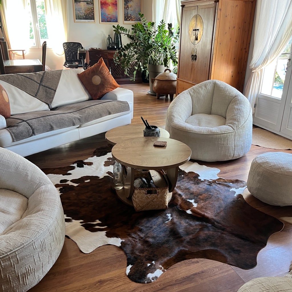 Peau de vache brun foncé et blanche sous deux tables basses en bois, dans un salon lumineux avec des sièges blancs.